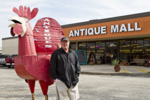 Man with big paper Mache red rooster in front of antique mall